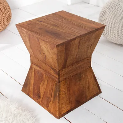 A wooden side table with a unique hourglass shape, placed on a white wooden floor with a fluffy white rug and a woven pouf in the background.