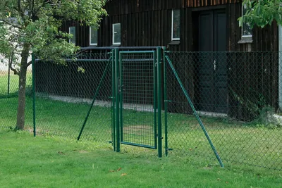 A green metal garden gate is shown in an outdoor setting, with a wooden building and a chain-link fence in the background.