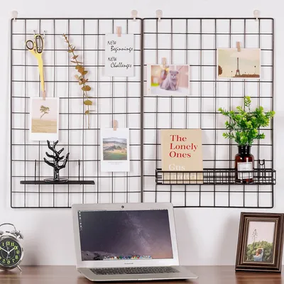 A grid wall display with various items attached, including photos, a plant, a book, and scissors, with a laptop and a framed photo on the desk below.