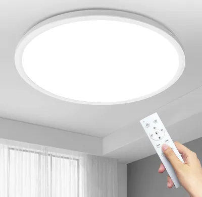 A hand holds a remote control in front of a white, round LED ceiling light mounted on a white ceiling. Sheer curtains are visible in the background.