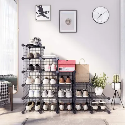 A black wire cube shoe rack filled with various shoes, boxes, and a hat, placed in a room with artwork and a clock on the wall.