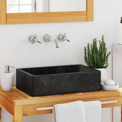 A bathroom sink setup with a black rectangular basin on a wooden vanity, featuring wall-mounted faucets, a soap dispenser, and a plant.