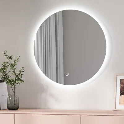 A round LED mirror with a white light ring is mounted on a light gray wall above a light pink dresser. A vase with green leaves is on the left side of the dresser.