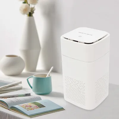 A white air purifier sits on a table next to a mug and some books, with vases in the background.