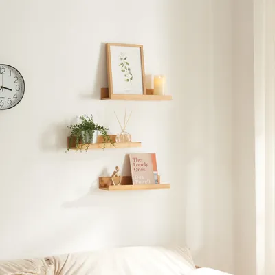 Three wooden shelves mounted on a white wall, decorated with a framed picture, a plant, a candle, a diffuser, and a book.