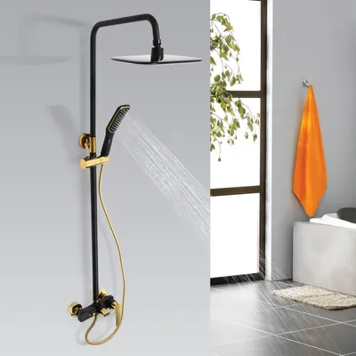 A black and gold shower system with a rainfall showerhead and handheld sprayer is shown against a light gray wall. Water is spraying from the handheld showerhead. A window with green foliage is visible in the background, along with an orange towel hanging