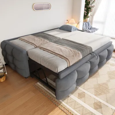 A gray sofa bed is shown in a room with wooden floors and a rug. The sofa bed is partially extended, revealing a metal frame underneath. There are blankets and a pillow on the sofa bed.