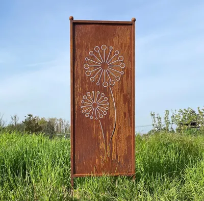 A rusted metal garden screen with a dandelion motif stands in tall green grass under a blue sky.