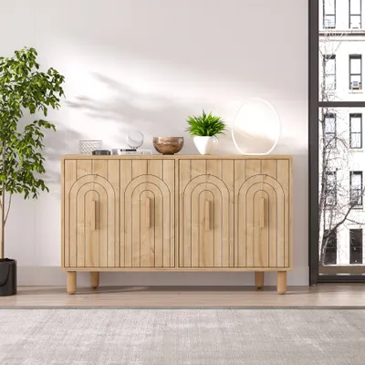 A light wood sideboard with decorative arch patterns on the doors, placed in a bright room with a large window on the right and a potted plant on the left. Decorative items and a plant are on top of the sideboard.