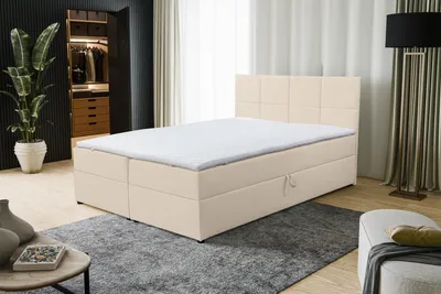 A beige boxspring bed with a white mattress topper is shown in a bedroom setting. The bed has storage drawers. A large plant is visible in the background.