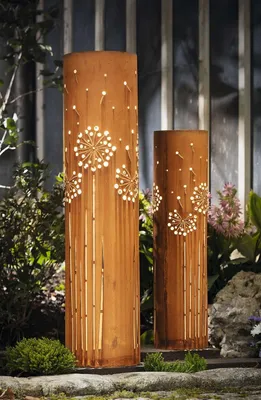 Two metal decorative columns with cut-out dandelion designs are illuminated by internal lights, placed in a garden setting with plants and rocks.