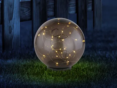 A solar-powered garden light ball with LEDs sits on grass in front of a wooden fence at dusk.