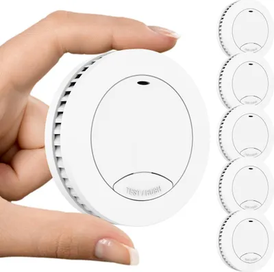A hand is holding a white smoke detector, with a stack of five more smoke detectors visible on the right side of the image.