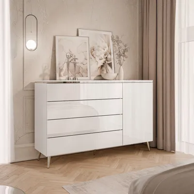 A modern white dresser with silver legs and chrome accents, placed in a bedroom setting with artwork and curtains.