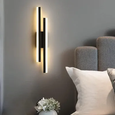 A modern wall lamp is mounted on a gray wall next to a bed with pillows.