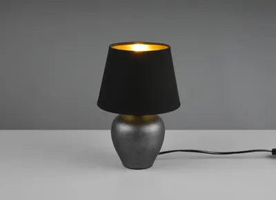 A small table lamp with a black fabric shade and a textured gray ceramic base sits on a white surface against a gray background.