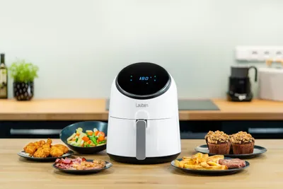 An air fryer sits on a kitchen counter surrounded by various foods, including fried chicken, vegetables, fries, and muffins.