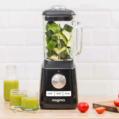 A black Magimix blender filled with chopped green vegetables, placed on a wooden countertop with two glasses of green smoothie and some tomatoes.