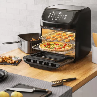 An air fryer oven with food inside is on a wooden countertop, with kitchen items around it.