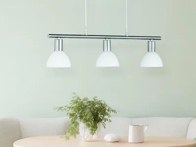 A three-light pendant lamp with white glass shades hangs above a table with a plant and a mug.