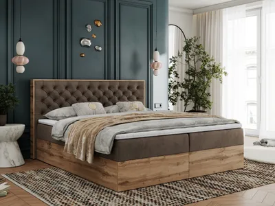 A bedroom scene featuring a large boxspring bed with a tufted headboard, decorative pillows, and a throw blanket. The room has dark teal walls, modern pendant lights, and a large plant.