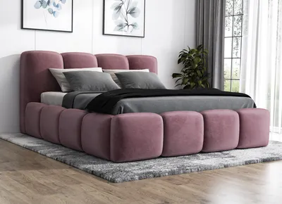 A pink upholstered bed with a headboard and footboard, set up in a bedroom with artwork on the wall and a plant in the background.