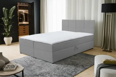 A bedroom scene featuring a gray boxspring bed with a white mattress and headboard. A large plant is visible in the background, along with a closet and a floor lamp.