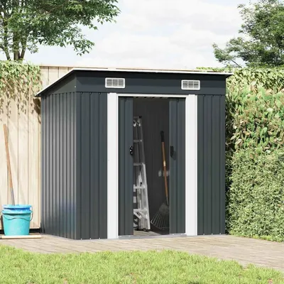 A dark gray metal garden shed with open doors, revealing a ladder and rake inside. It is situated on a wooden deck next to a wooden fence and a green hedge, with grass in the foreground.