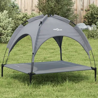 A gray dog bed with a canopy is set up on a grassy lawn in front of a wooden fence and some bushes.