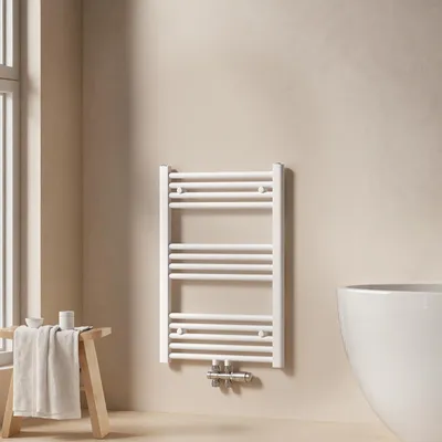 A white towel radiator is mounted on a light beige wall in a bathroom. To the left, a wooden stool with a folded towel and some small containers is placed near a window. To the right, a portion of a white bathtub is visible.