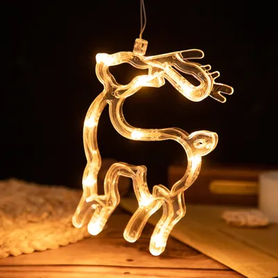 A close-up of a clear LED reindeer decoration with warm white lights, hanging against a dark background and resting on a wooden surface.