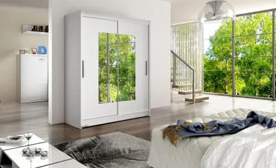 A modern bedroom with a large white wardrobe with mirrored doors reflecting a lush green outdoor scene. A bed with white bedding and a blue throw is in the foreground.