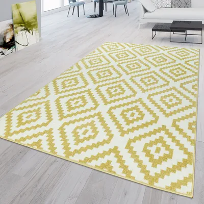 A rug with a geometric pattern is laid out on a light-colored wooden floor in a modern living room setting. The rug is yellow and white with a diamond pattern. In the background, there is a white sofa, a black coffee table, and dining chairs around a table