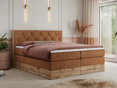 A bedroom scene featuring a king-size boxspring bed with a tufted headboard and wooden base. The bed is dressed with bedding in muted tones. Decorative wall lights and a vase with dried flowers add to the ambiance. A bedside table with a plant is visible o