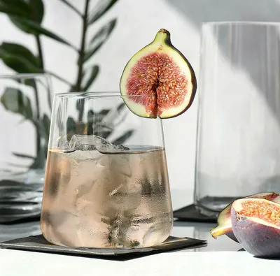 A close-up of a glass tumbler filled with a pinkish beverage and ice, garnished with a fig slice. Another glass and figs are visible in the background.