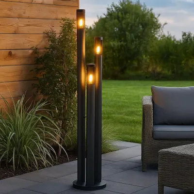 An outdoor patio scene at dusk with a set of three modern black LED bollard lights illuminating the area. A wicker chair and ottoman are visible to the right, and a wooden fence and lush greenery form the background.