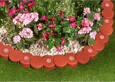 A garden bed border made of interlocking brown segments with white circular inserts, surrounding pink and red flowers and green foliage, set against a green lawn.