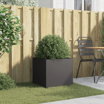 A black planter with a green bush sits on a lawn in front of a wooden fence. A chair and table are visible to the right.