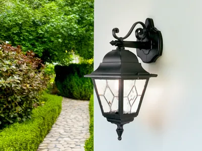 An outdoor wall lantern is mounted on a white wall next to a garden path and lush greenery.