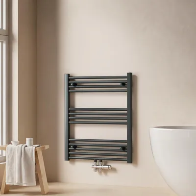A dark gray towel radiator is mounted on a light beige wall in a bathroom setting. A wooden stool with a folded towel and a small white bowl is placed to the left of the radiator. Part of a white bathtub is visible on the right.