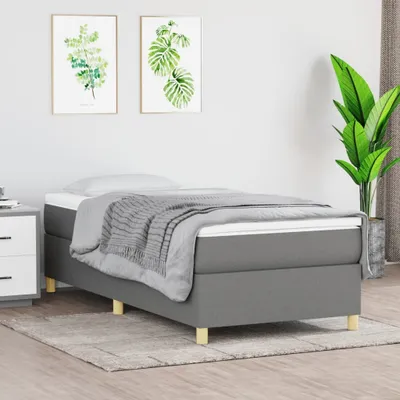 A single bed with gray fabric upholstery and wooden legs, neatly made with a white mattress, gray bedding, and a gray throw blanket. Two framed botanical prints hang on the wall behind the bed, and a large potted plant is visible on the right side of the i
