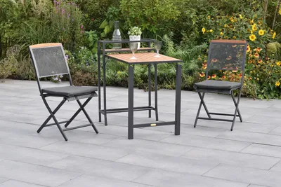 A patio set with two chairs and a table is set up on a paved patio, surrounded by lush greenery and flowers.