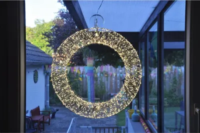 A lit wreath hangs in front of a window overlooking a backyard.