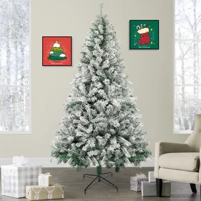 A flocked artificial Christmas tree is displayed in a room with two framed Christmas-themed pictures on the wall and wrapped gifts at its base.