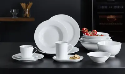 A collection of white ceramic dishes and bowls are arranged on a dark countertop, with a kitchen oven visible in the background.
