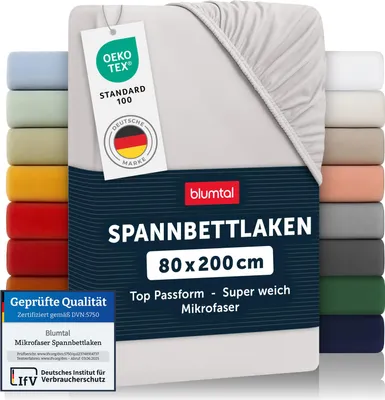 A light gray fitted sheet is shown folded and partially unfolded, with a stack of other colored fitted sheets visible on the left and right sides. Several labels and certifications are attached to the sheet.