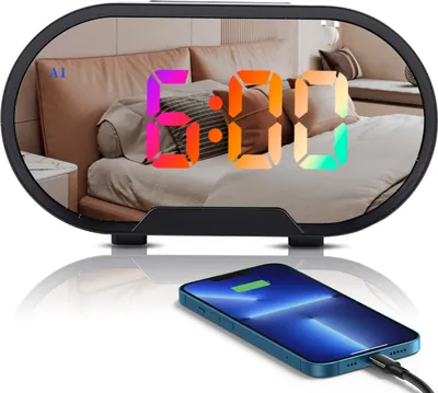 A digital alarm clock with a mirrored surface reflecting a bedroom scene, and a smartphone charging in front of it.