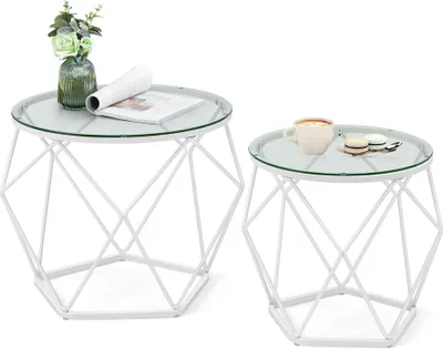 Two white geometric side tables with glass tops, one with a vase of flowers and a magazine, the other with a cup and macarons.
