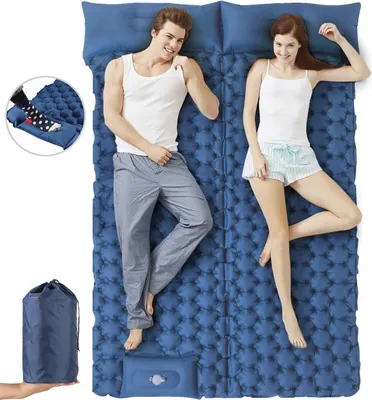 A couple lying on a blue inflatable sleeping mat, smiling at the camera. The mat is designed for two people and includes an integrated pillow. A rolled-up sleeping mat in a carrying bag is shown in the lower left corner.