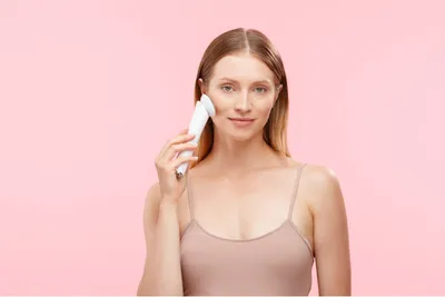 A woman is holding a white facial cleansing device to her cheek against a pink background.